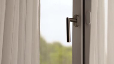 Açık bir pencerenin yakın çekim görüntüsü gösterişli bir kulp ve dalgalı perdeler, dışarıdaki canlı yeşillikten hafif bir esinti ve güneş ışığı davet ederek barışçıl bir atmosfer yaratıyor..