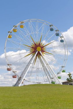 Büyük, renkli bir dönme dolap, kabarık bulutlu parlak mavi bir gökyüzüne karşı dimdik ayakta duruyor. Geniş yeşil bir parkta yer alan park, ziyaretçileri manzaranın tadını çıkarmaya davet ediyor..