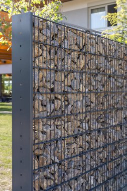Taşlarla dolu metal tel kafeslerden yapılmış eşsiz bir gabion çiti manikürlü bir avluda duruyor. Bu çağdaş tasarım dış mekanlara yenilikçi bir dokunuş ekler.