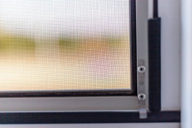 Ilık güneş ışığı, kısmen açık bir pencere ekranından geçer ve dışarıdaki yeşilliğin yumuşak odak noktasını gösterir. Davetkar ambiyans yakınlardaki huzurlu bir doğal ortamı işaret ediyor..