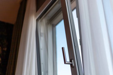 Kısmen açık bir pencere, yumuşak perdelerin güneş ışığını çerçevelediği açık mavi gökyüzüne bir bakış sağlar. İçerisi rahat ve davetkar, rahatlamak için mükemmel..