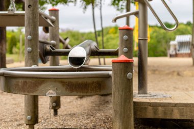 Bir oyun parkında dairesel bir havzanın üzerine uzanan gümüş piposu olan bir su özelliği sergileniyor. Gri gökyüzünün altındaki yeşil bir parkta tahta direkler ve oyun ekipmanları görülebiliyor..