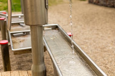 Metal bir su pompası çocuklar için tasarlanmış uzun bir kanala temiz su dağıtıyor. Sahne, arka planda kumlu bir yüzey ve yeşillikle eğlenceli..