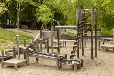 Çocukların oyun parkında, gün ışığında ağaçlarla çevrili çimenli bir alanda fiziksel aktivite ve keşif için tasarlanmış çeşitli metal oyun ekipmanları sergileniyor..