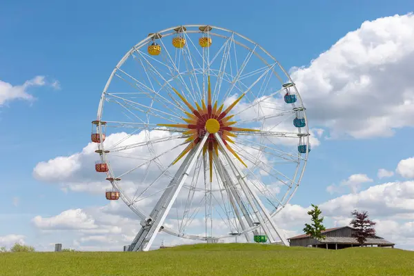 Renkli gondolları olan büyük bir dönme dolap tüylü bulutlarla dolu canlı mavi bir gökyüzüne çarpar. Çevreleyen çimenler yemyeşil ve yemyeşil, canlı bir atmosferi gösteriyor..