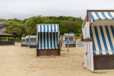 Mavi ve beyaz çizgili plaj sandalyeleri bir kıyı bölgesinin kumlu sahilinde düzenlenmiştir. Gökyüzü bulutlu, sakin bir atmosfer yaratıyor..