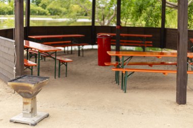 Bu açık hava buluşma yerinde barınağın altına yerleştirilmiş turuncu piknik masaları sergileniyor. Yakınlarda bir ızgara ve huzurlu bir park ortamında bir çöp tenekesi var..