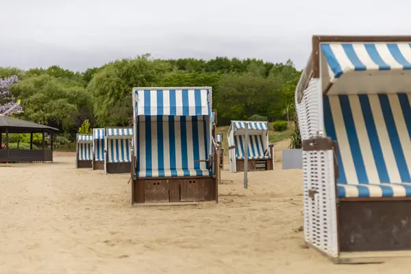 Mavi ve beyaz çizgili plaj sandalyeleri bir kıyı bölgesinin kumlu sahilinde düzenlenmiştir. Gökyüzü bulutlu, sakin bir atmosfer yaratıyor..