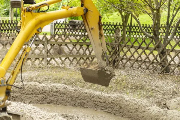 Sarı bir kazıcı, ahşap bir çitle çevrili bir inşaat alanında toprak taşıma görevini yerine getiriyor. Makineler temiz gökyüzünün altında toprak dolu bir kova kaldırır..