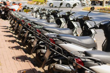 Kaldırımda düzgün park edilmiş uzun bir scooter kuyruğu var. Parlak, güneşli bir gün, arkalarındaki işlek sokakla çelişen araçların krom ve renklerini vurguluyor..