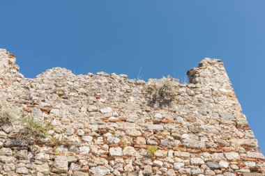 Yıpranmış taş duvar canlı mavi bir gökyüzüne karşı duruyor, tarihi önemini ve mimari detaylarını gözler önüne seriyor. Doğa bu yapının bazı kısımlarını geri almaya başladı..