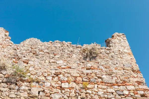 Yıpranmış taş duvar canlı mavi bir gökyüzüne karşı duruyor, tarihi önemini ve mimari detaylarını gözler önüne seriyor. Doğa bu yapının bazı kısımlarını geri almaya başladı..