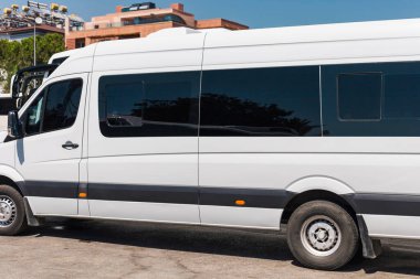 Güneşli bir günde büyük beyaz bir minibüs, parlak bir tasarım ve renkli camlar sergiliyor, kentsel bir alanda çağdaş mimari ile çevrili..