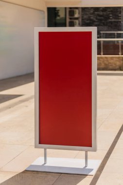 Açık havada, metalin tabanında boş kırmızı bir tabela var. Parlak tasarım, açık renkli taş döşeme ve yakınlardaki binalarla çelişiyor..