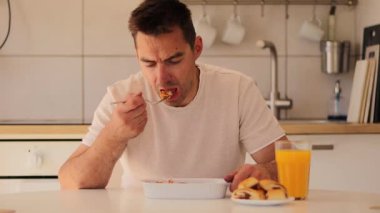 Bir adam açık mutfak masasında oturmuş, ferahlatıcı portakal suyunu yudumlarken doyurucu bir yemek yiyor. Atmosfer parlak ve davetkar, modern bir mutfak alanı sergiliyor.