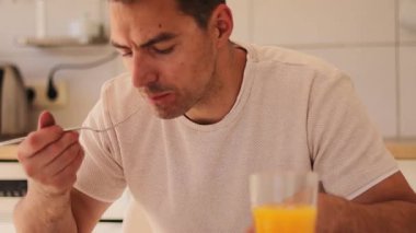 Bir adam açık mutfak masasında oturmuş, ferahlatıcı portakal suyunu yudumlarken doyurucu bir yemek yiyor. Atmosfer parlak ve davetkar, modern bir mutfak alanı sergiliyor.