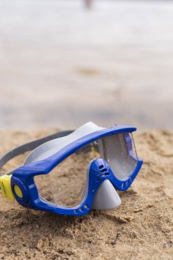 Mavi bir şnorkelli maske sıcak kumun üzerinde bir sonraki su altı macerasını bekliyor. Yumuşak dalgalar, parlak bir gökyüzünün altında huzur dolu bir sahil atmosferi oluşturur..