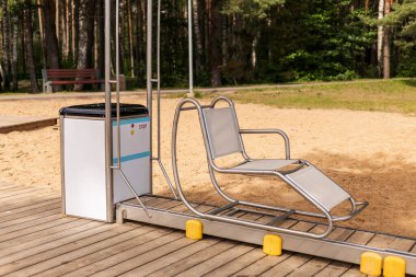 Tahta bir platformda oturan sürgülü sandalye engelli ziyaretçilerin suya girmesine yardım etmeye hazır. Kurulum kumsalda, ağaçlar ve kumlu zemin ile çevrili..