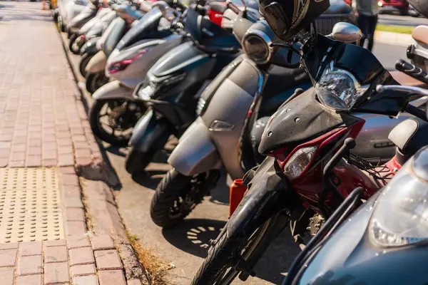 Açık mavi gökyüzünün altında kalabalık bir şehir caddesi boyunca sayısız motosiklet ve scooter park edilmiştir. Sahne, şehir hayatının canlılığını yakalıyor..