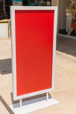 A tall sign with a bright red background is placed on the sidewalk. It stands empty, ready for announcements or promotions. The setting is a lively urban area on a sunny day.