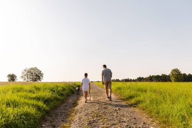 Bir adam ve küçük oğlu uzun otlarla çevrili bir çakıl yolunda yürürler. Köpek mutlu bir şekilde yol gösterirken güneş tepemizde parlıyor..