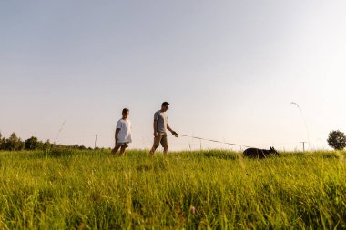Bir adam ve küçük oğlu yeşil bir arazide tasma tutarken sakince yürüyorlar. Dost canlısı köpekleri onlara eşlik eder, sıcak güneşin tadını çıkarırlar..