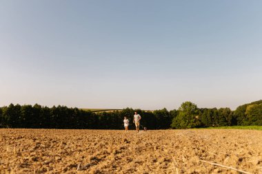 Bir adam ve küçük oğlu açık alanda mutlu mesut yürüyorlar. Açık mavi gökyüzünün altındaki açık hava manzarasını keşfederken köpekleri de onları yakından takip ediyor..