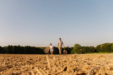 Bir adam ve oğlu, köpekleriyle birlikte kuru bir tarlada birlikte yürüyorlar. Gökyüzü berrak, sıcak bir güneş parlıyor ve huzurlu bir atmosfer yaratıyor..