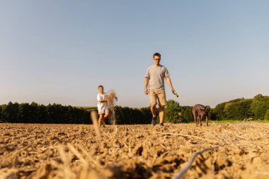 Bir adam ve oğlu büyük, açık bir alanda birlikte yürüyorlar. Yanlarında neşeli bir köpek var, sıcak havanın ve parlak güneş ışığının tadını çıkarıyorlar. Çevreleyen ağaçlar huzurlu atmosfere katılıyor..