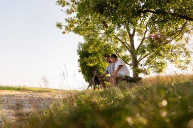 Sakin bir parkta, bir adam oğlu ve köpeğinin yanındaki bankta oturup sıcak bir öğleden sonranın tadını çıkarıyor. Yeşillik ve yumuşak güneş ışığıyla çevrili sessiz bir anı paylaşıyorlar..