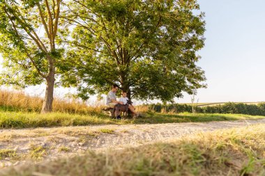 Baba ve oğul bir ağacın altında oturup kırsalda güneşli bir günün tadını çıkarıyorlar. Dost canlısı köpekleri çimenli yolda uzanmış, aile bağlarını ön plana çıkarıyor..