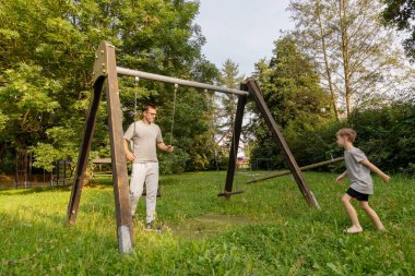 Bir baba ve küçük oğlu parkta salıncakta birlikte oynuyorlar. Ağaçlarla çevrili çimenli alanda eğlenirken gülüp kaynaşırlar..