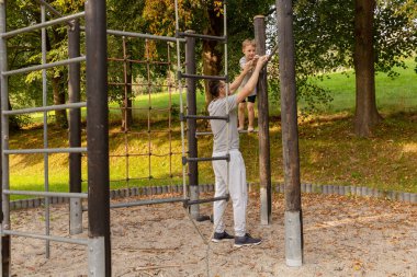 Etrafı ağaçlarla çevrili sakin bir oyun alanında bir baba, küçük oğlunun bir binaya tırmanmasına yardım eder. Kahkaha ve neşeyi paylaşıyorlar, oyun ve keşif yoluyla bağlarını güçlendiriyorlar..