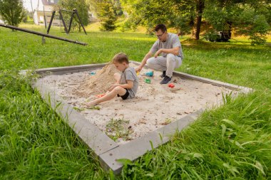 Bir baba ve küçük oğlu kum havuzunda birlikte oynuyorlar, kumda eğlenceli şekiller yaratıyorlar. Parlak ve neşeli bir günde etrafları ağaçlarla ve çimlerle çevrilidir..
