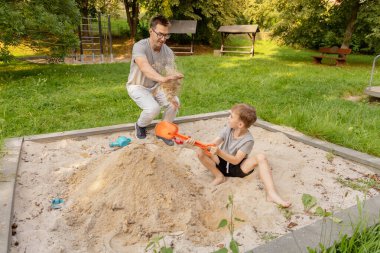 Bir baba ve oğlu ağaçlar ve çimlerle çevrili bir kum havuzunda oynarken eğleniyorlar. Baba kumu kaldırırken çocuk parlak turuncu bir kürekle kazıyor ve bağlarını gösteriyor..