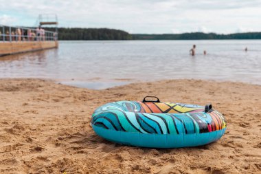 Canlı bir şamandıra halkası bir gölün yakınındaki kumlu sahilde duruyor. Arka planda, insanlar açık bir gökyüzünün altında yüzerken ve dinlenirken görülebilir..
