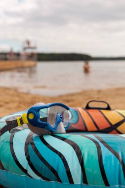 Canlı bir şişme yüzük kumlu sahilde duruyor, şnorkelle dalış takımlarıyla eşleştirilmiş. Yakınlarda, sakin bir göl insanların suyun tadını çıkarırken gri bulutları yansıtıyor ve sakin bir atmosfer yaratıyor..