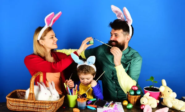 Easter family traditions. Mother, father and son painting eggs for holidays. Happy family in bunny ears painting Easter eggs together. Happy family preparing for Easter. Rabbits family on Easter