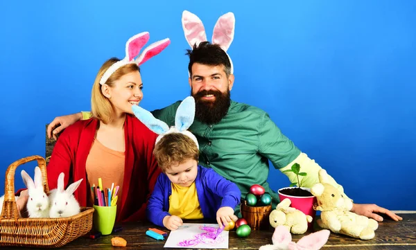 Easter family traditions. Happy parents and adorable boy in bunny ears sitting together at table with colorful painted eggs. Spring holidays. Mother, father and son preparing for Easter holiday