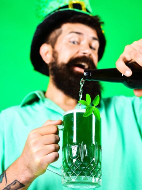 Aziz Patrick Günü. Bardağa yeşil bira dolduran mutlu adam. Yeşil şapkalı sakallı leprikon ve bir bardak birada yonca. Bir kupa yeşil biraya odaklan. İrlanda gelenekleri. Bar partisi kutlaması.