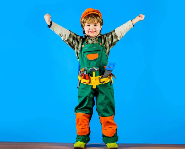 Smiling kid boy in safety helmet and toolbelt raised hands. Little repairman, builder or construction worker with tools for building. Child in builder uniform playing with toy tools for repair