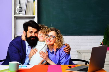 Okula dönelim. Evde eğitim. İlkokuldan bir çocuk. Anne baba ve oğul birlikte okuyorlar. Eğitimsel süreç. Okul ailesi. Mutlu erkek ve bayan öğretmenler küçük okul çocuğuna sarılıyorlar.