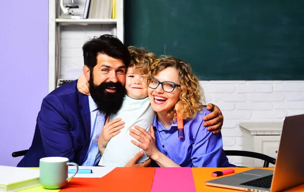Birinci sınıftaki küçük okul çocuğu. İlkokuldan bir çocuk. Eğitimsel süreç. Okul ailesi. Anne baba ve oğul birlikte okuyorlar. Mutlu erkek ve bayan öğretmenler küçük okul çocuğuna sarılıyorlar.