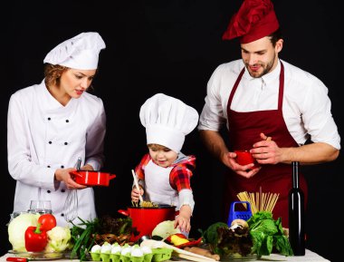 Aşçı üniformalı mutlu bir aile evde yemek pişiriyor. Aile mutfakta birlikte lezzetli yemekler hazırlıyor. Çocuklu aileler vejetaryen yemeği hazırlıyorlar. Anne ve baba küçük oğluna yemek yapmayı öğretiyor.