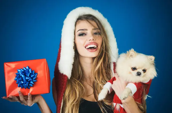 Christmas woman with little dog and gift box. Smiling girl in fur hat with Pomeranian Spitz in Santa Claus costume and Christmas present. Christmas and New year celebration. Pet and winter holidays