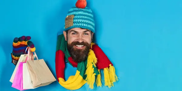 Smiling bearded man in winter clothes with shopping bags looking through paper hole. Handsome male in colourful hat, scarf and gloves with small gift bags. Gifts and presents. Season sales. Discount