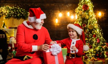 Mutlu Noeller ve mutlu yıllar. Noel Baba kılığındaki gülümseyen küçük çocuk ve Noel hediyeli Noel Baba. Aile tatilleri. Noel Baba ve küçük Noel Baba Noel için dekore edilmiş odada