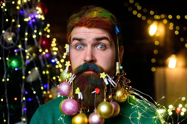 Christmas barbershop advertising. Bearded guy with decorated beard ready for Christmas or New Year party. Surprised man with colorful hair and beard with Christmas balls in beard. Closeup portrait