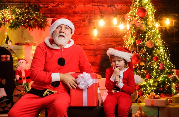 Thoughtful child boy and bearded man in Santa costume in room decorated for Christmas. New year present for children. Santa Claus and little Santa helper at home with Christmas gift. Family holidays