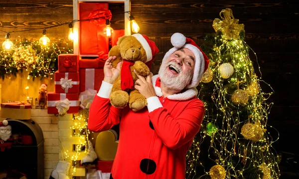 Christmas and New Year holidays. Christmas decoration. Happy Santa Claus with teddy bear on shoulder in room decorated for Christmas. Bearded man in Santa suit with toy teddy bear near Christmas tree